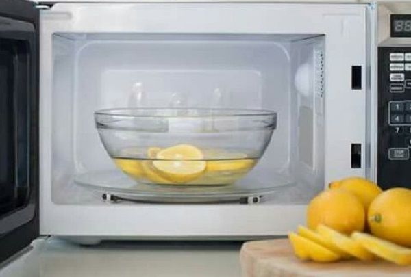 Simple Hacks to Clean Your House - Bowl half-full of white vinegar and lemon slices inside an open microwave with lemon slices on top of a wooden table in front of the microwave